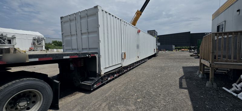 Long, wide modular unit chained down on a lowboy deck at a project site, with a crane boom behind it