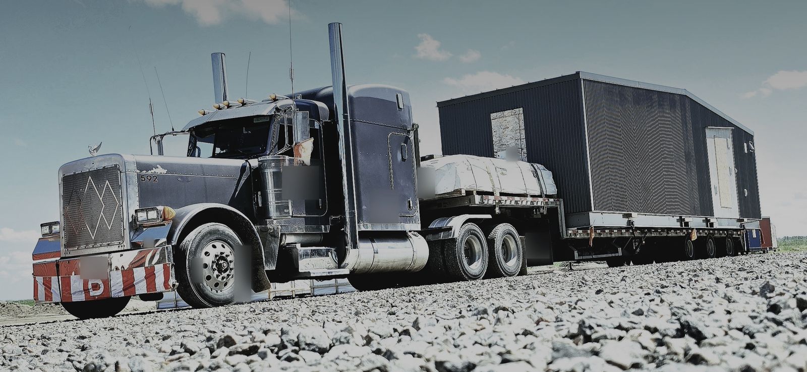 Long-nose truck with tall stacks pulling a multi-axle lowboy loaded with a wide modular building on a gravel pad
