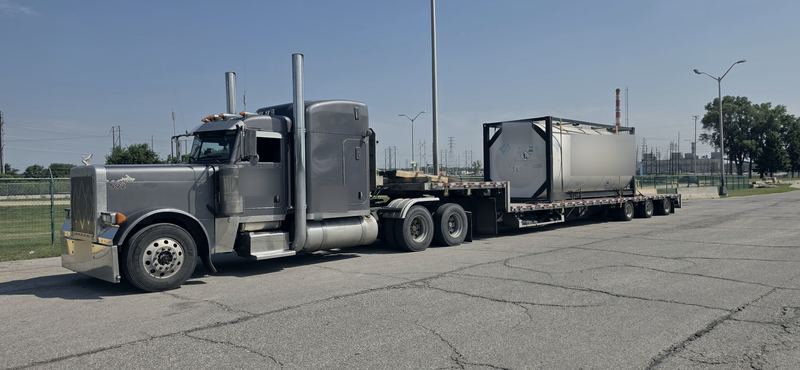 Grey Peterbilt 379 pulling a step deck trailer with a tank container, ready for a cross-border run