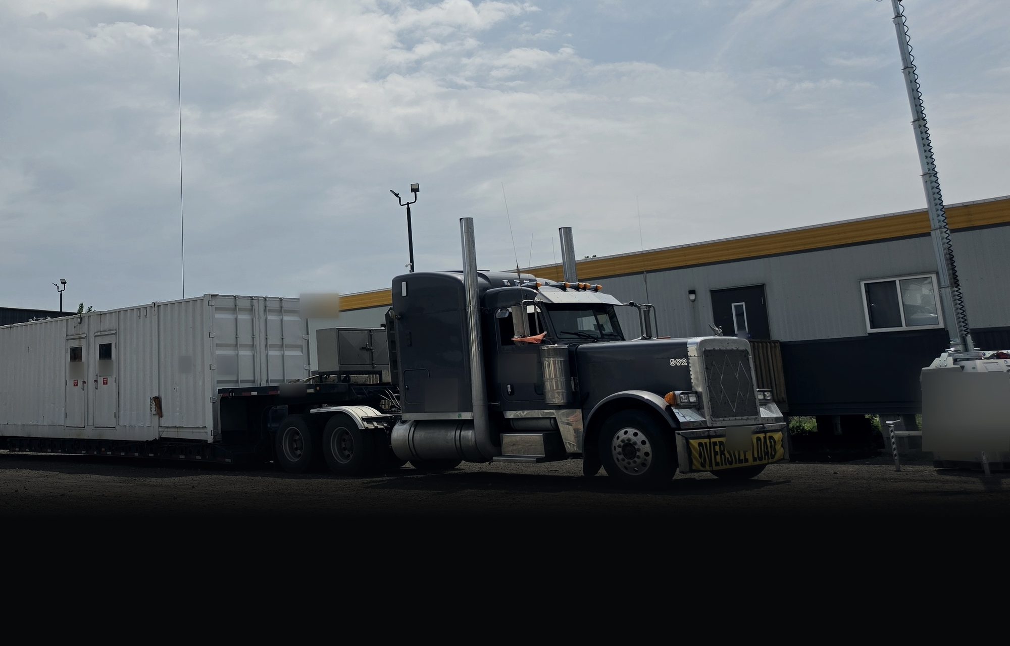 Long-nose Peterbilt 379 with an oversize-load banner beside a lowboy as a crane loads a long modular unit