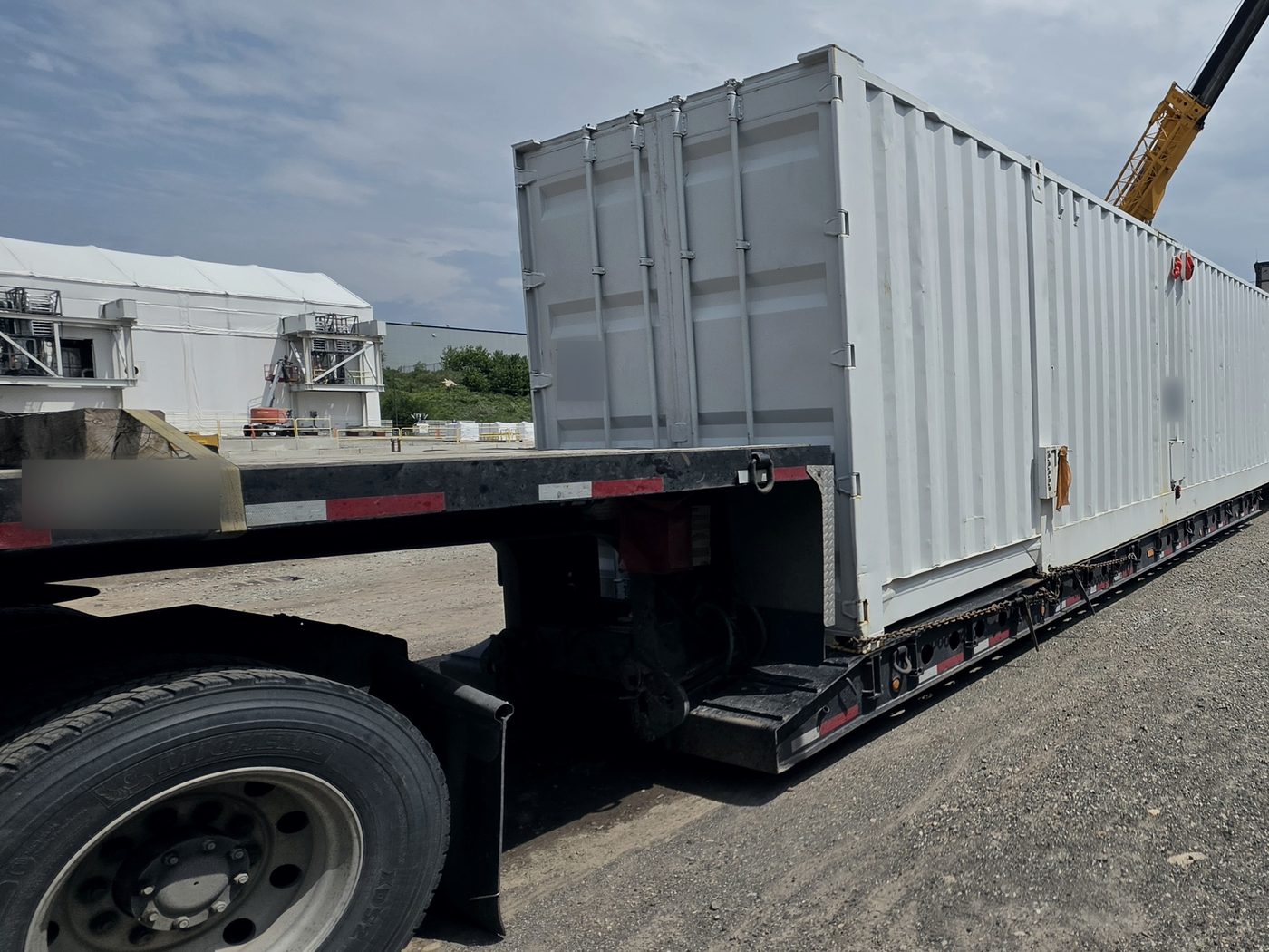 Long modular camp unit loaded on a lowboy deck at a project site, with a crane boom behind it